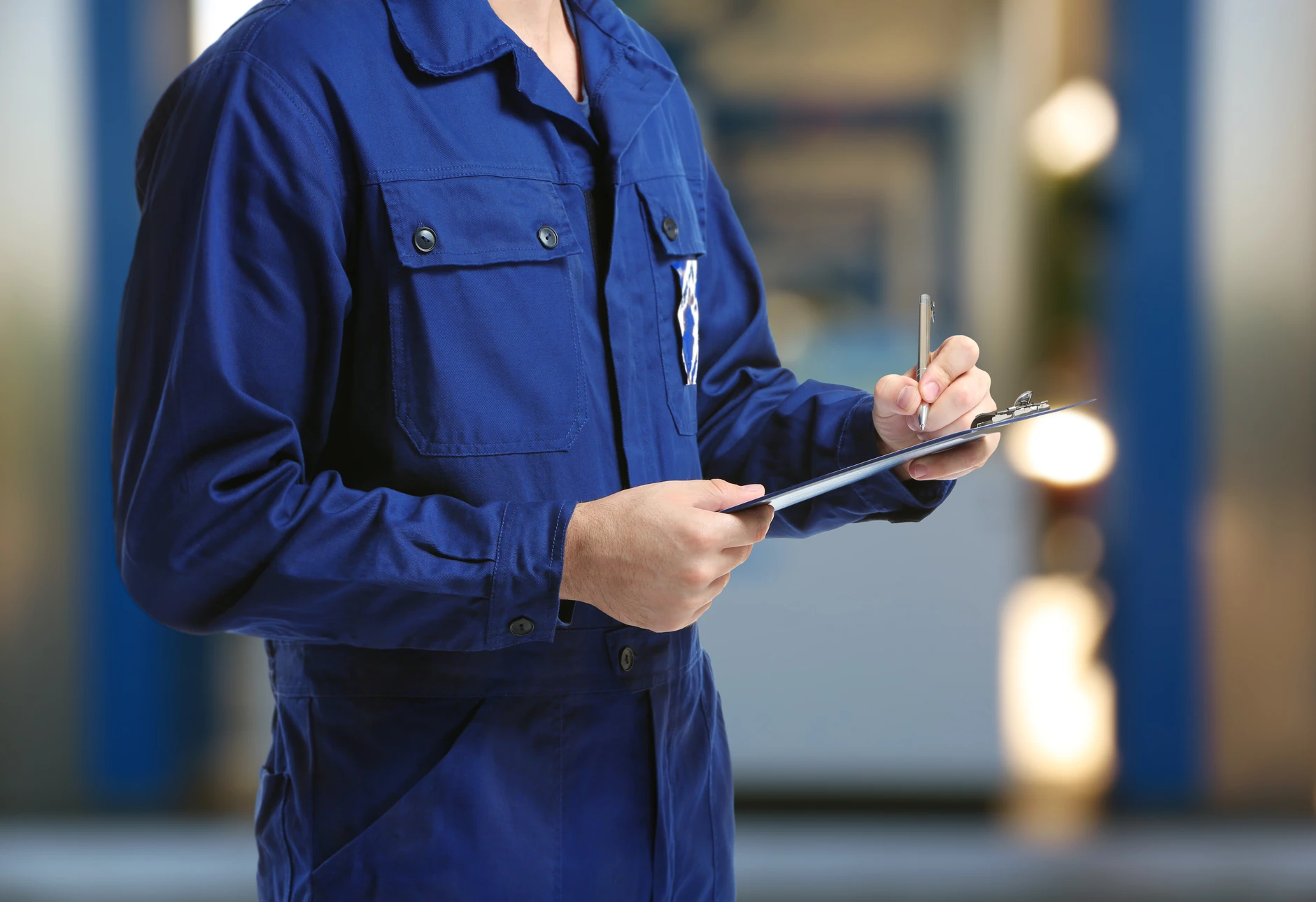 Professionnel en combinaison de travail bleue tenant un bloc-notes et un stylo, effectuant une inspection ou un contrôle qualité dans un environnement industriel extérieur avec équipements flous en arrière-plan