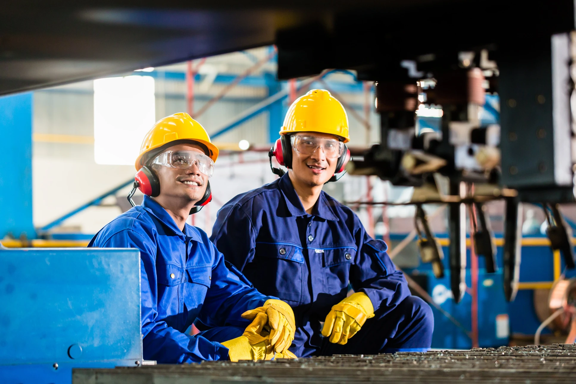 Deux ouvriers en combinaisons bleues assis dans un environnement industriel, portant casques jaunes, lunettes de protection et casques antibruit rouges