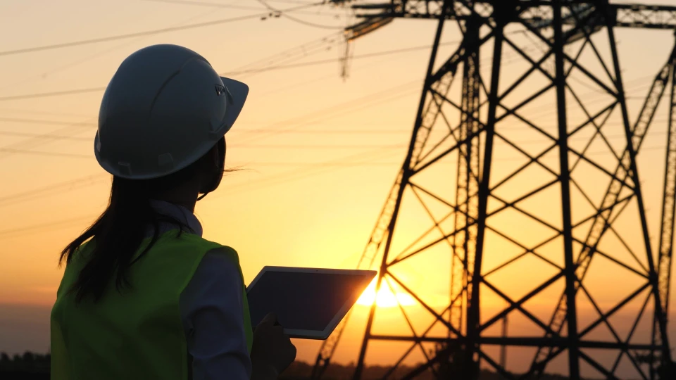 Scène extérieure au coucher du soleil avec ingénieure en tenue de sécurité verte et casque blanc consultant une tablette numérique près d'un pylône électrique haute tension