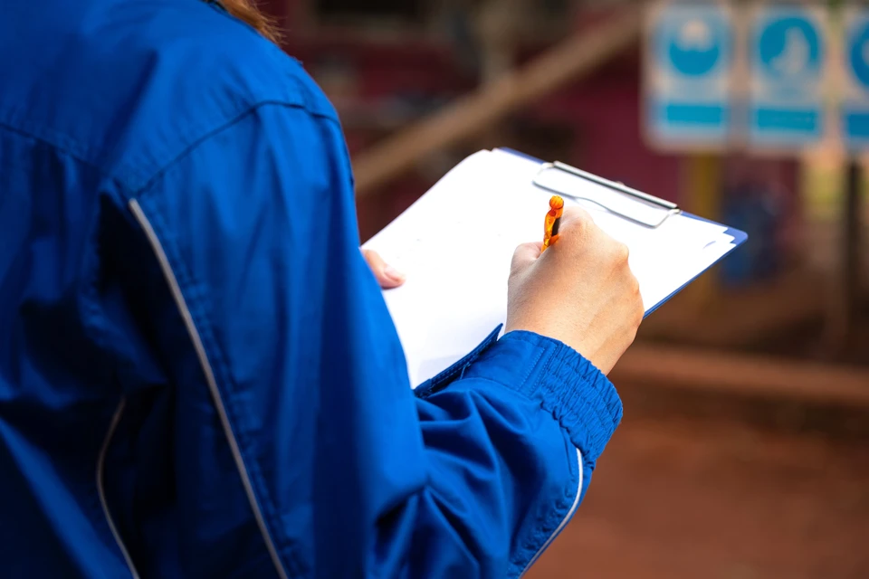 Homme en combinaison bleue de travail effectuant une inspection ou audit sur site industriel, tenant un porte-documents avec formulaires et stylo orange