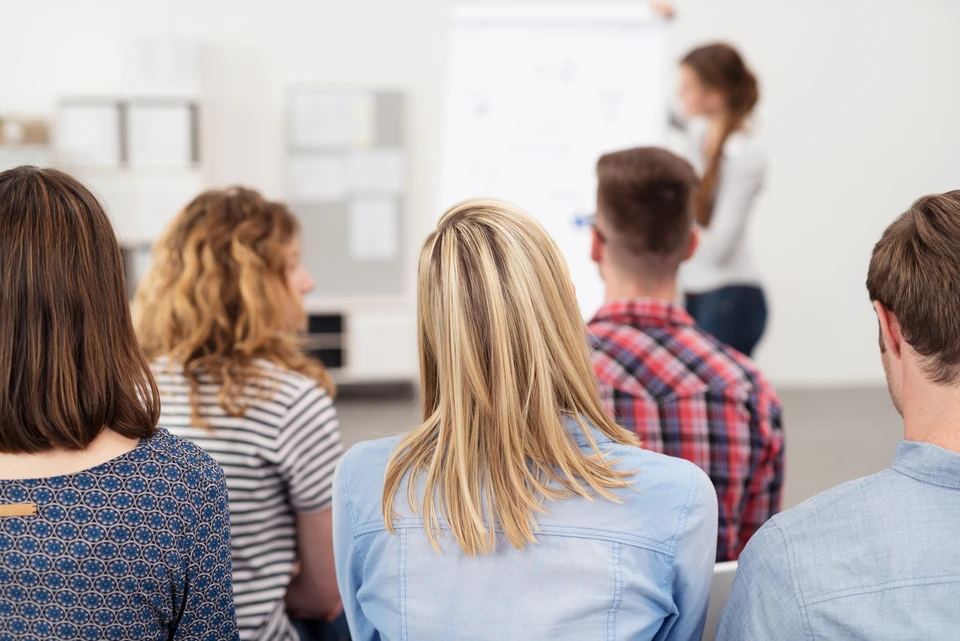 Salle de formation moderne avec groupe d'adultes assis face à une formatrice debout devant tableau blanc