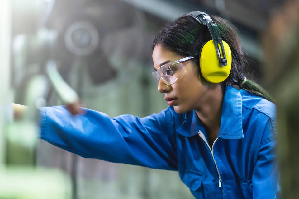 Femme ingénieure en combinaison bleue travaillant dans un atelier industriel, portant casque antibruit jaune et lunettes de protection transparentes
