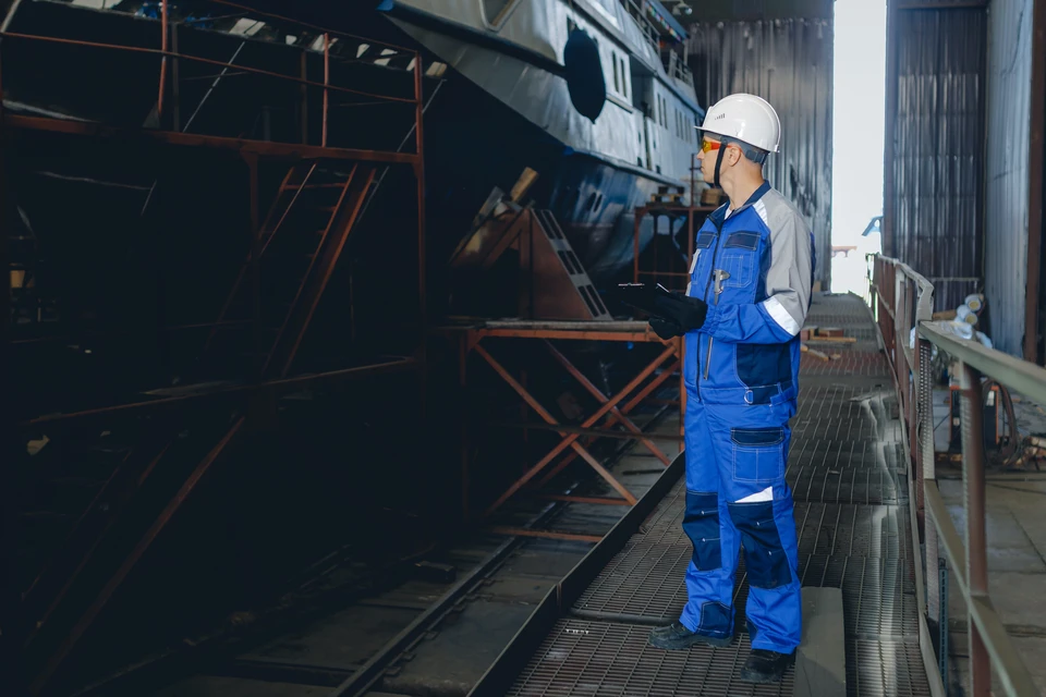 Professionnel en tenue de protection (combinaison, casque de sécurité, masque) effectuant une inspection ou une mesure de radioactivité avec un dosimètre ou un équipement de détection dans un environnement nucléaire ou naval sécurisé (installation portuaire, zone contrôlée avec signalétique radioactive visible en arrière-plan). L'accent doit être mis sur la rigueur technique et la conformité réglementaire du travail en milieu à risque radiologique.