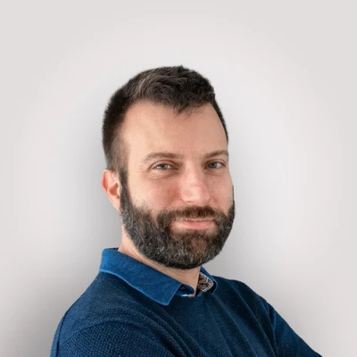 Portrait professionnel d'un homme souriant en intérieur, veste grise sur pull bordeaux, barbe soignée, cheveux bruns coiffés, regard direct et bienveillant, expression confiante et chaleureuse